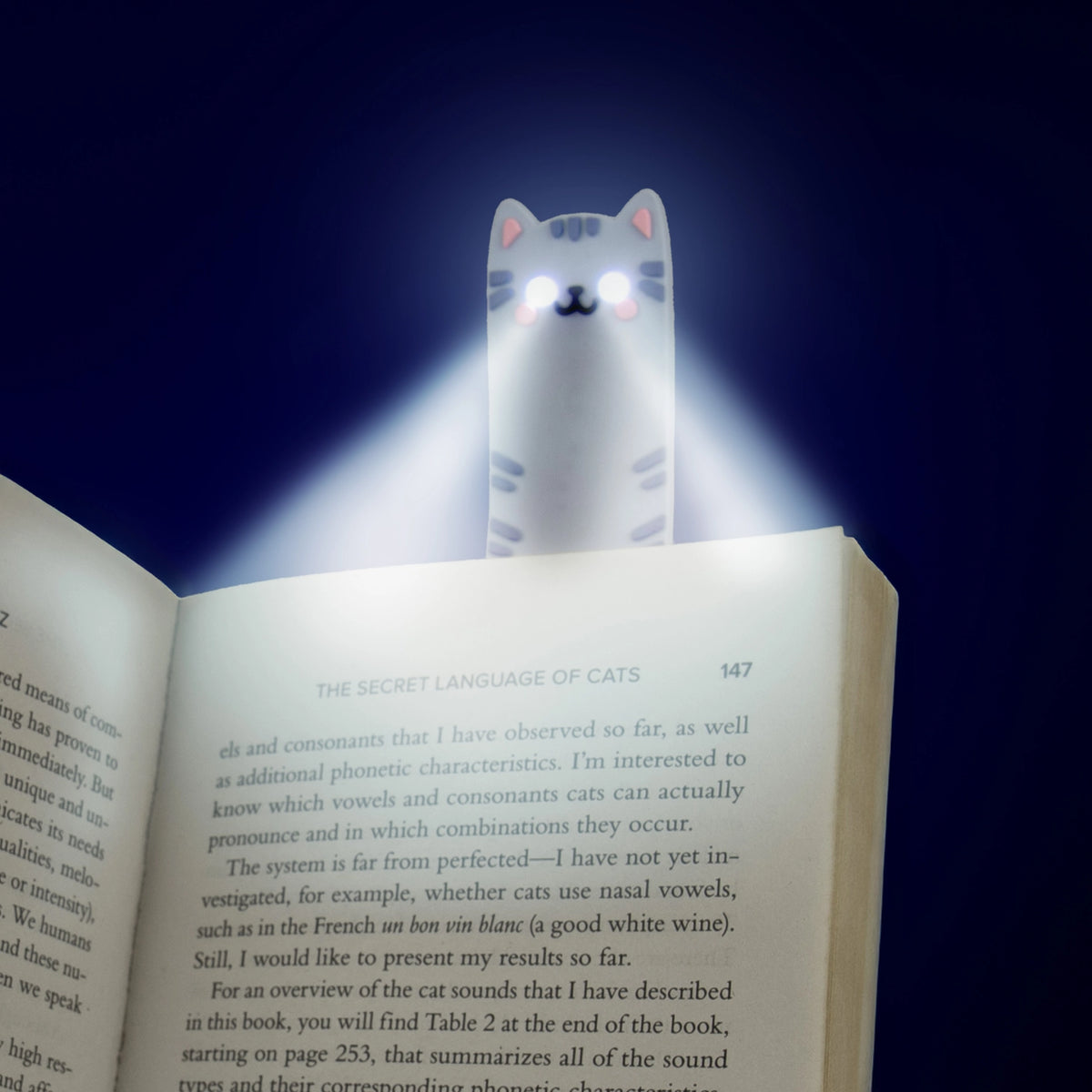 Cat reading light glowing with bright white LED beams in a dark room, clipped to the top of an open book for nighttime reading.