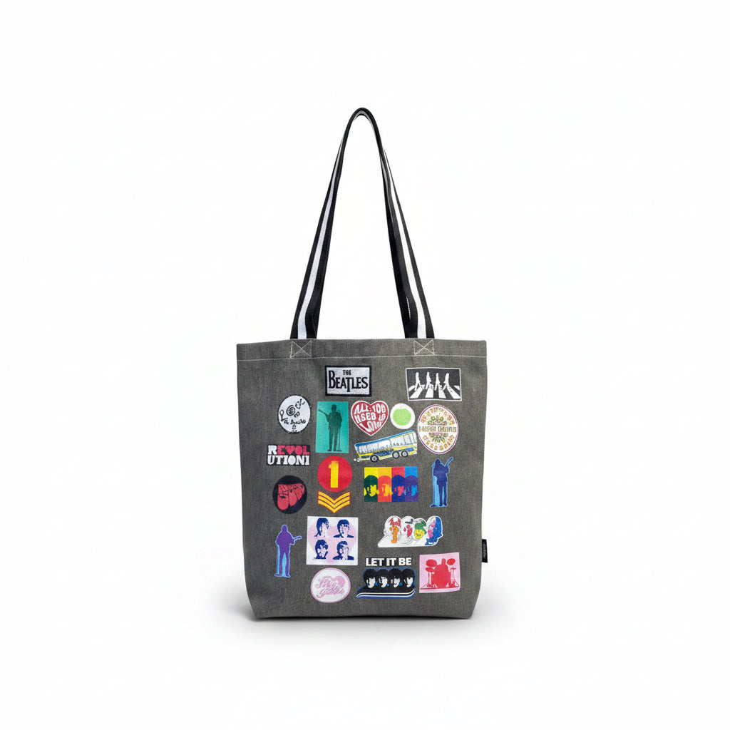 Person wearing grey Beatles tote bag with colourful printed patch designs and black-and-white straps, next to a yellow submarine model.
