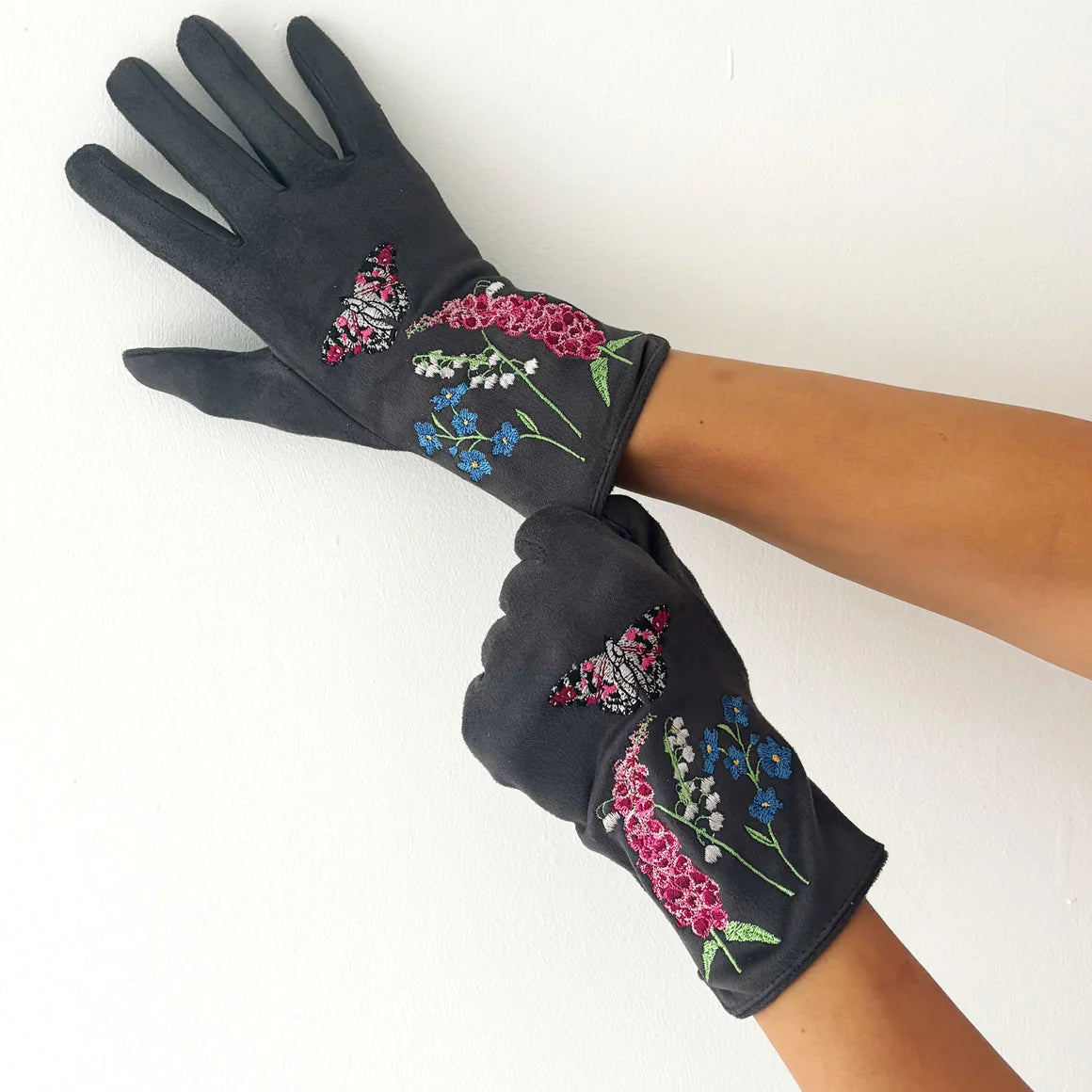 Person wearing grey suedette gloves embroidered with pink butterflies, foxgloves, lily of the valley, and blue cornflowers, shown with arms crossed against a white background.