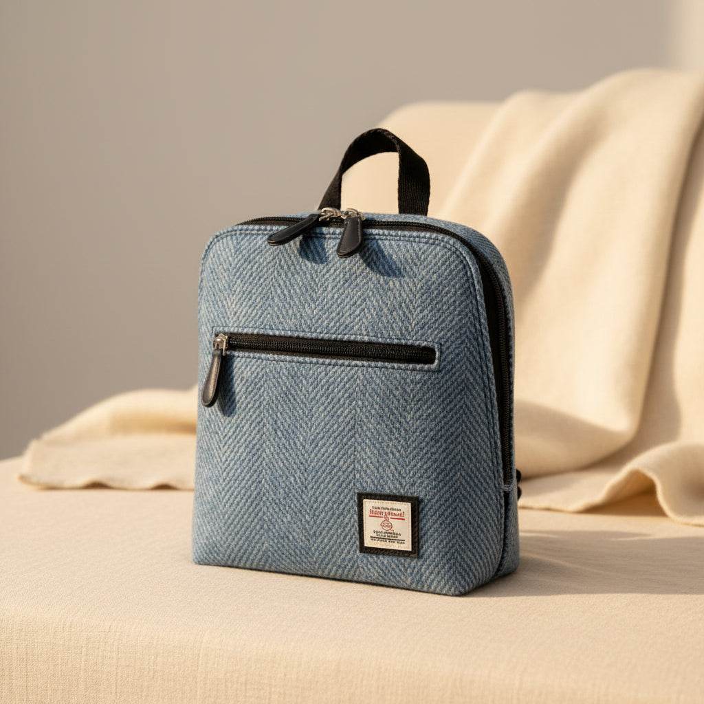 A blue herringbone tweed travel pouch or mini backpack with black trim, a top handle, a front zippered pocket, and a small brand patch on the lower right, shown on a white background.