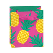 Mini pineapple gift bag with bright yellow fruit and green leaves on a bold pink background, featuring matching pink grosgrain ribbon handles and a gift tag. Made from recyclable FSC-certified paper.