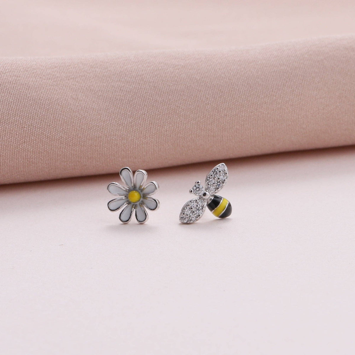 Close-up of enamel bee and daisy stud earrings on dusty pink fabric