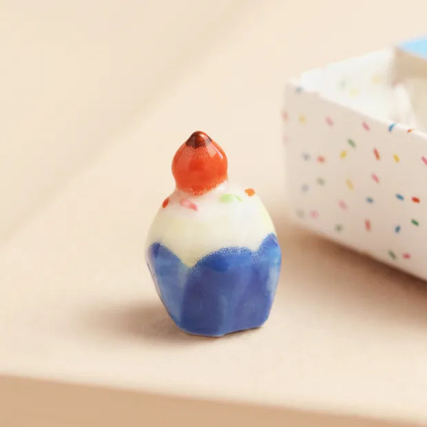 Ceramic cupcake token with blue case and cherry on top beside confetti matchbox on cream background
